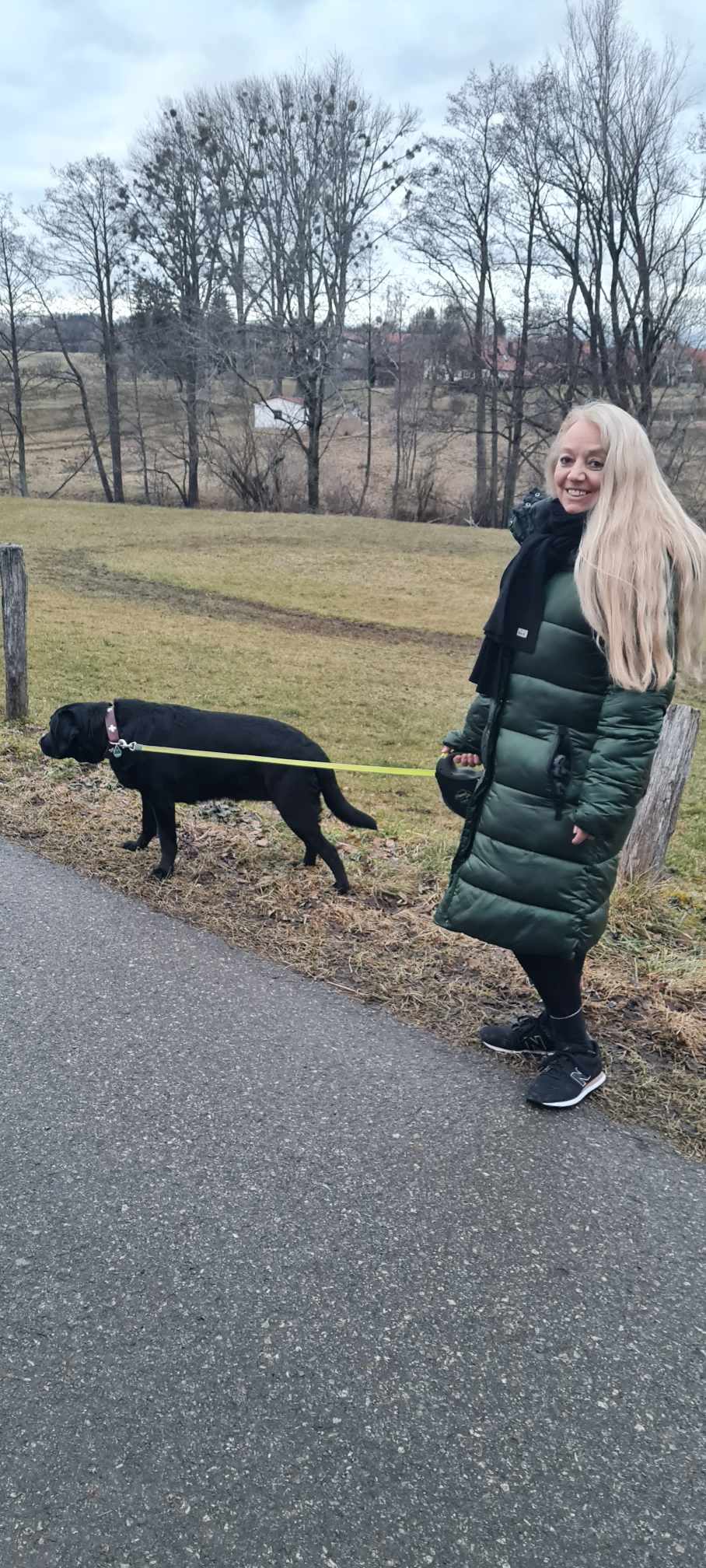 Ältere Frau mit langen, blonden Haaren und ihrem schwarzen Hund an der Leine.